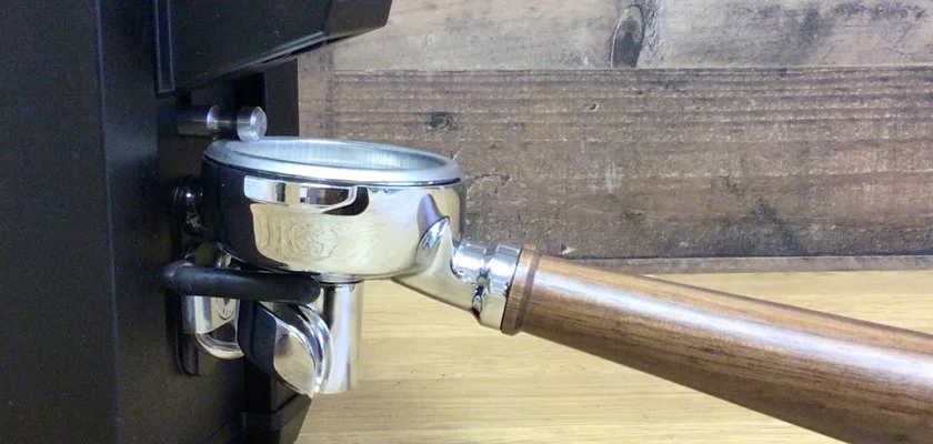 A portafilter with a wooden handle is attached to an espresso machine, ready for brewing. Nearby on the wooden surface sits the Macap Leo 55 Espresso Coffee Grinder, completing this inviting coffee setup. © 2025 1st-line.com