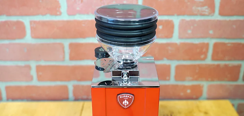 A red and chrome Eureka Mignon Zero Espresso Coffee Grinder with a clear bean hopper and black lid sits on a wooden surface against a red brick wall, displaying the Eureka logo on the front. © 2025 1st-line.com