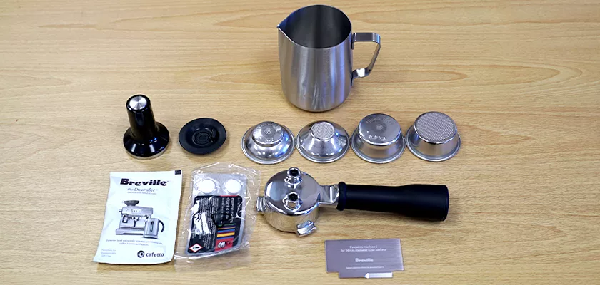 A set of accessories for the Breville Bambino Plus™ Espresso Machine on a wooden surface, featuring a metal milk frothing pitcher, tamper, filter baskets, portafilter, cleaning tool, user manual, and instruction cards. © 2025 1st-line.com