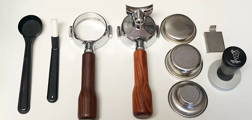 Accessories for the Bezzera Aria HX Special Edition Espresso Machine w/ Flow Control on a white surface, including a spoon, brush, two wooden-handled portafilters, filter baskets, tamper, and metal dosing tool for flow control. © 2025 1st-line.com