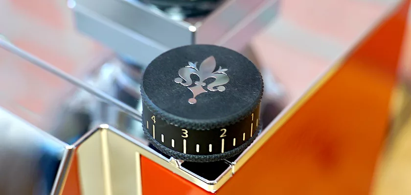Close-up of a black knob with a fleur-de-lis symbol and white numbers 0–5 on the Eureka Mignon Zero Espresso Coffee Grinder, mounted on a shiny metallic surface. © 2025 1st-line.com
