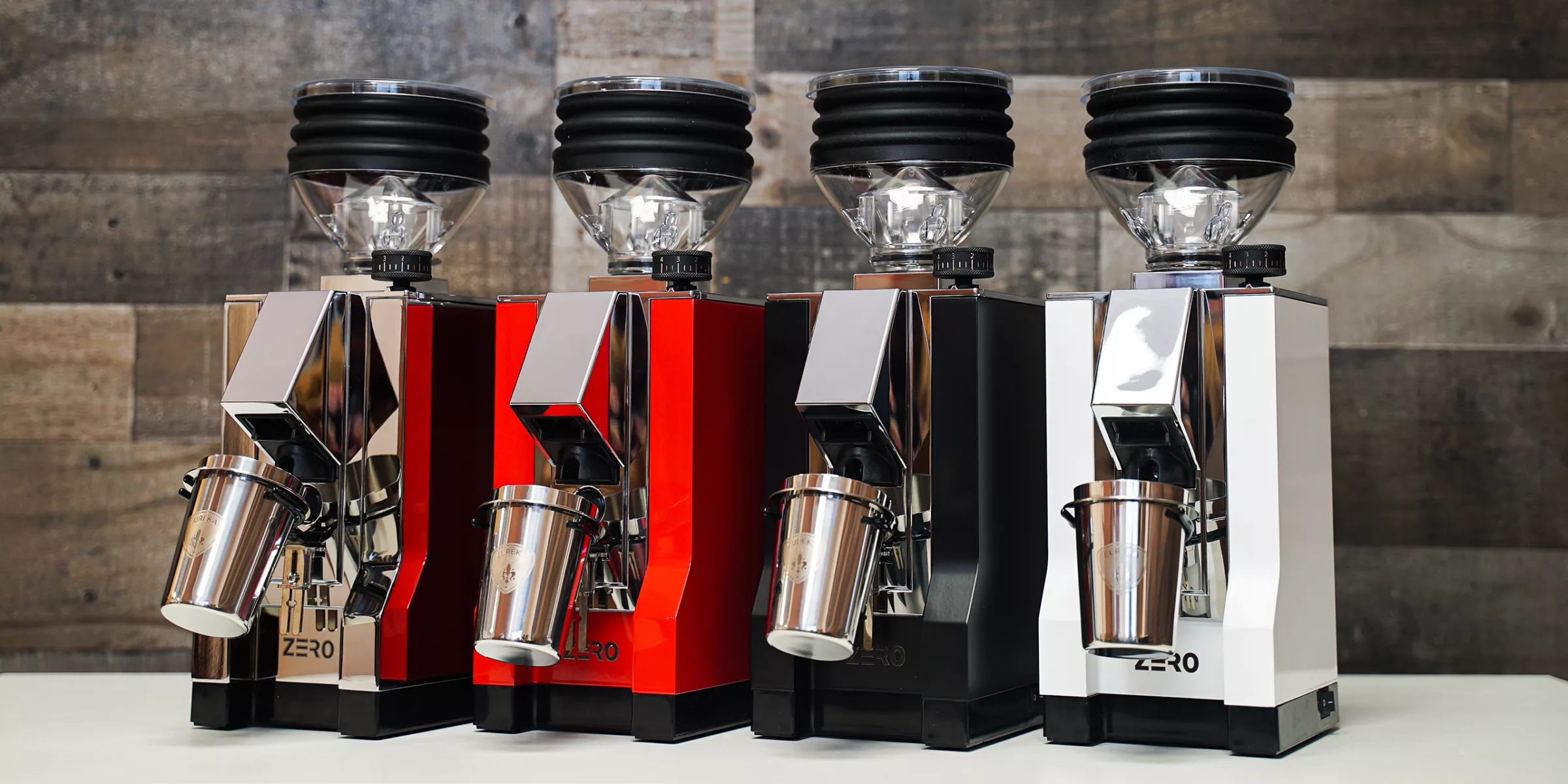 Four Eureka Mignon Zero Espresso Coffee Grinders in silver, red, black, and white are lined up on a white surface with a wooden background. Each grinder has a clear hopper on top and a metal cup attached in front. © 2025 1st-line.com
