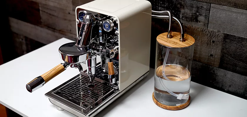 The DISCONTINUED ECM Puristika Espresso Machine, featuring wooden accents and a clear glass water reservoir with a wooden lid and handle, sits on a white surface against a textured dark wood wall. © 2025 1st-line.com