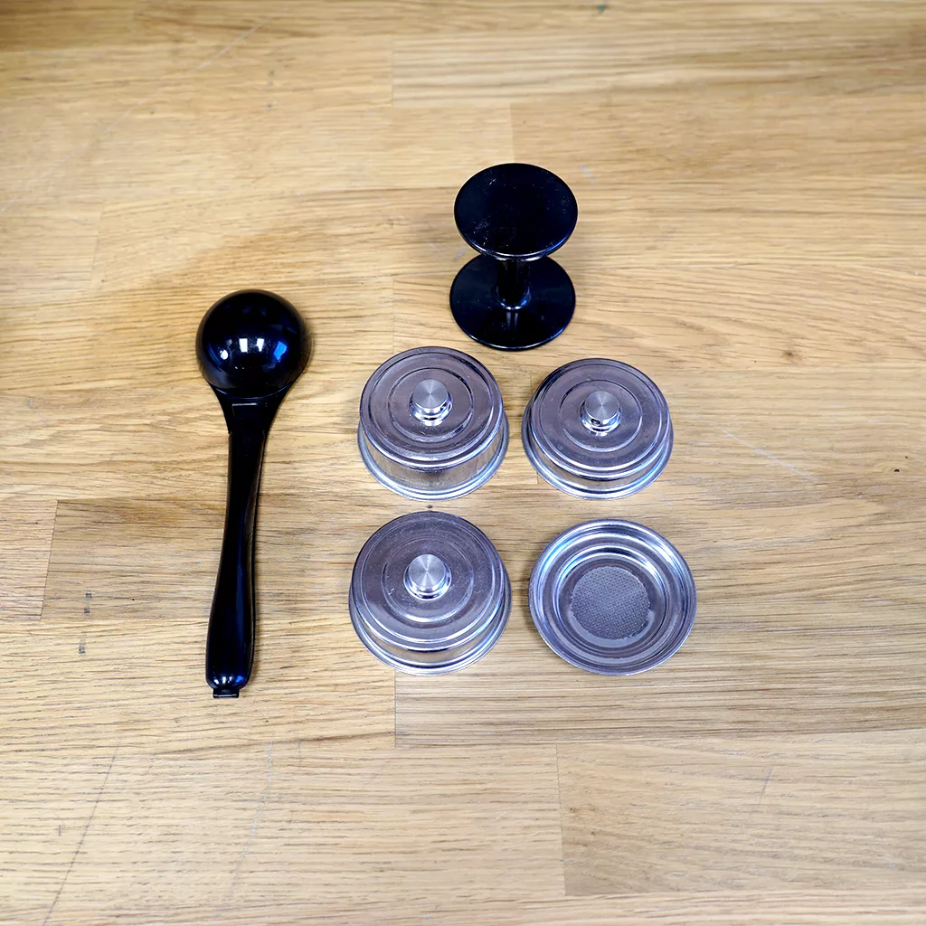 A black-handled coffee tamper, three espresso baskets, a mesh-bottom basket for Ascaso Duo Steel, and a dual-sided distributor sit on wood—ideal accessories for your (SOLD) SIDEWALK SALE - Ascaso Duo Steel Versatile Espresso Machine. © 2025 1st-line.com