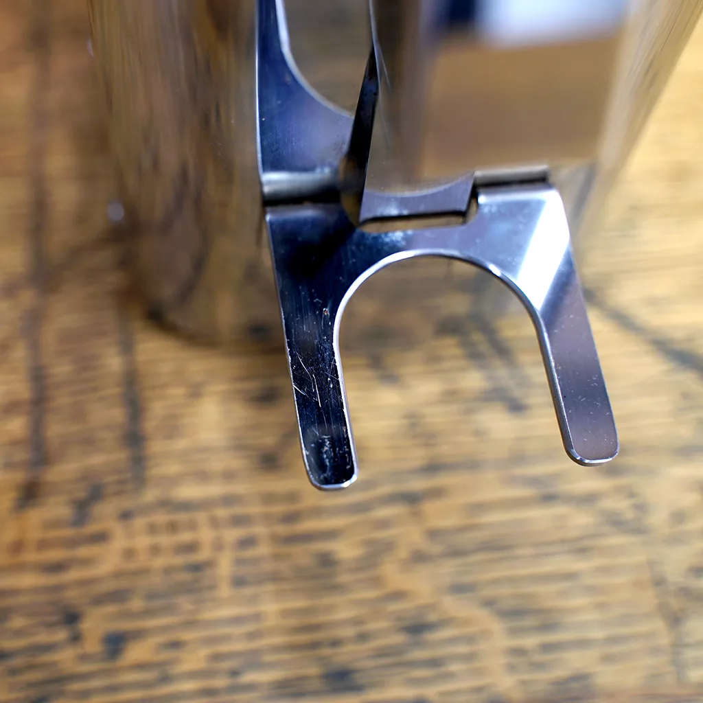 A close-up of a metal object with a U-shaped cutout, part of a SIDEWALK SALE - ECM C-Manuale 54 Espresso Grinder NOT FOR ESPRESSO (Returned and Refurbished) E209, resting on wood with visible grain. © 2025 1st-line.com