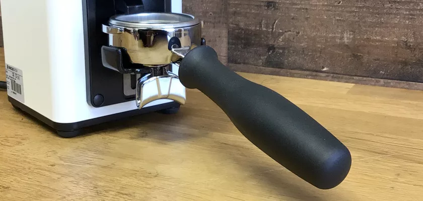 A portafilter with a black handle is attached to an espresso machine, next to a Rancilio Stile Espresso Coffee Grinder on a wooden countertop with a wooden wall in the background. © 2025 1st-line.com