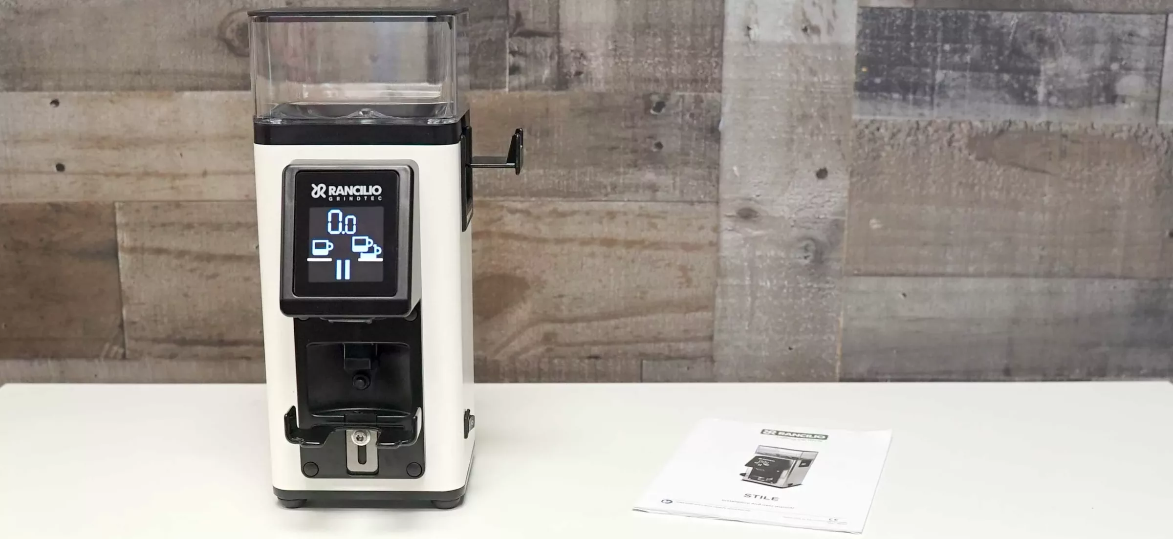 A Rancilio Stile Espresso Coffee Grinder in white, featuring a digital display, sits on a white surface beside its user manual, with a wooden textured wall in the background. © 2025 1st-line.com