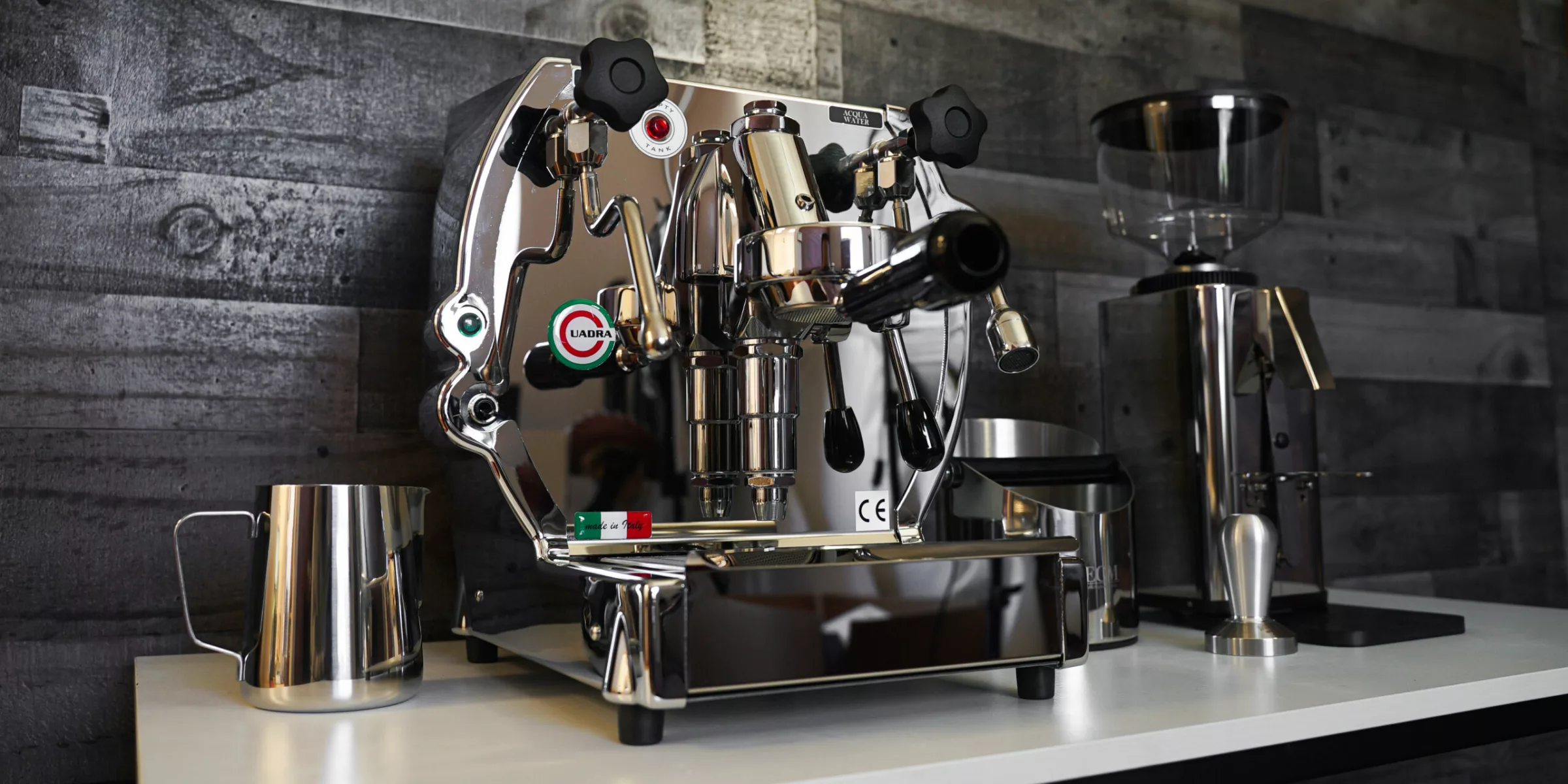 A shiny espresso machine with various knobs and levers sits on a counter beside a metal milk pitcher and quality grinders, all set against a dark, wooden tiled wall—perfect for complete package deals. © 2025 1st-line.com