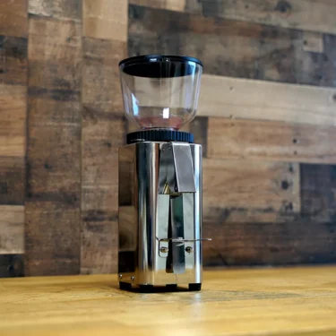 A SIDEWALK SALE - ECM C-Manuale 54 Espresso Grinder NOT FOR ESPRESSO (Returned and Refurbished) E209 with a transparent bean hopper is displayed on a wooden countertop in front of a rustic wooden wall. © 2025 1st-line.com