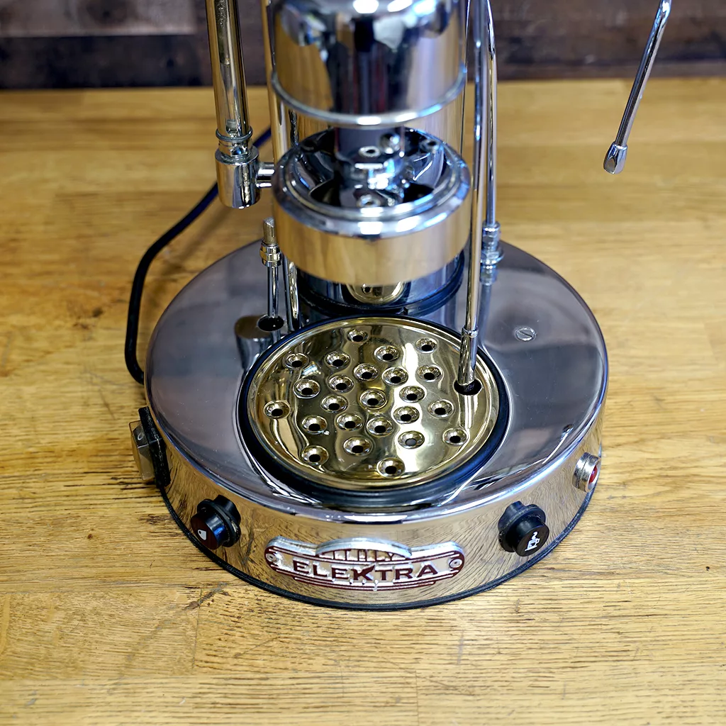 A SIDEWALK SALE - Elektra SXC Microcasa Semiautomatica Chrome Espresso Machine USED (E211) sits on a wooden countertop, featuring a brass drip tray with circular holes and black control knobs on the front. © 2025 1st-line.com