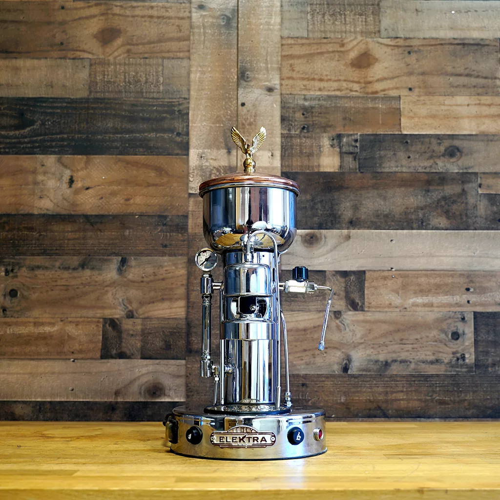 A USED Elektra SXC Microcasa Semiautomatica Chrome Espresso Machine (E211) from our SIDEWALK SALE, featuring a polished finish and eagle ornament, displayed on a wooden surface with a rustic wood wall background. © 2025 1st-line.com