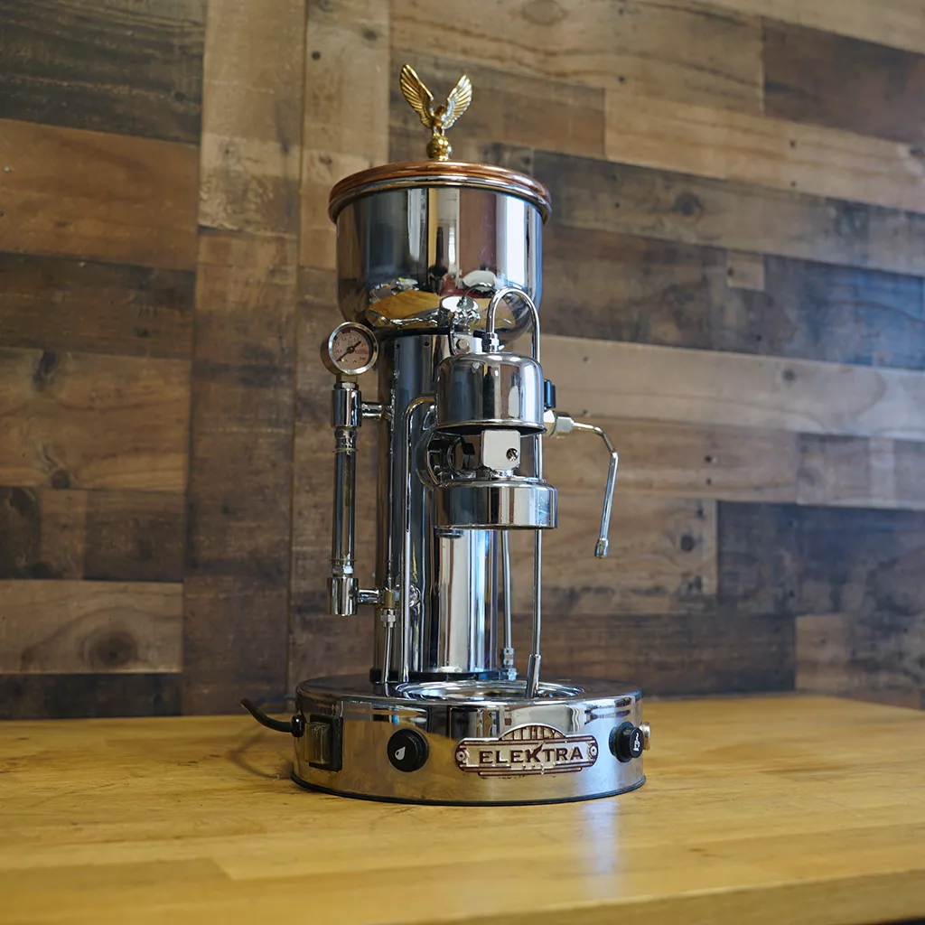 A USED Elektra SXC Microcasa Semiautomatica Chrome Espresso Machine (E211) from our sidewalk sale, featuring a polished metal finish and vintage design, is displayed on a wooden counter against a wood-paneled wall. © 2025 1st-line.com