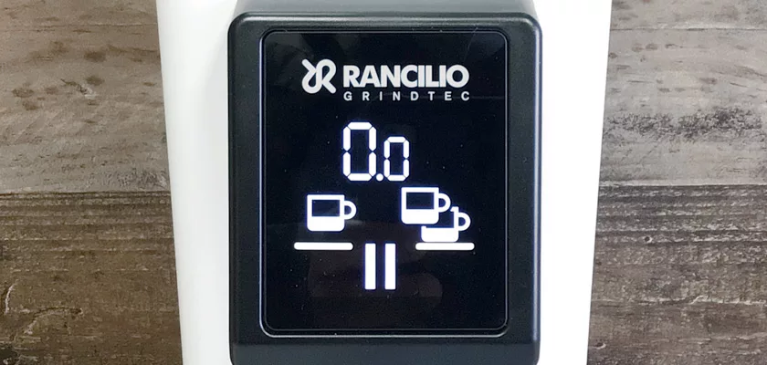 Close-up of the digital display on a Rancilio Stile Espresso Coffee Grinder, featuring single and double cup icons and a pause symbol, set against a wooden surface background. © 2025 1st-line.com