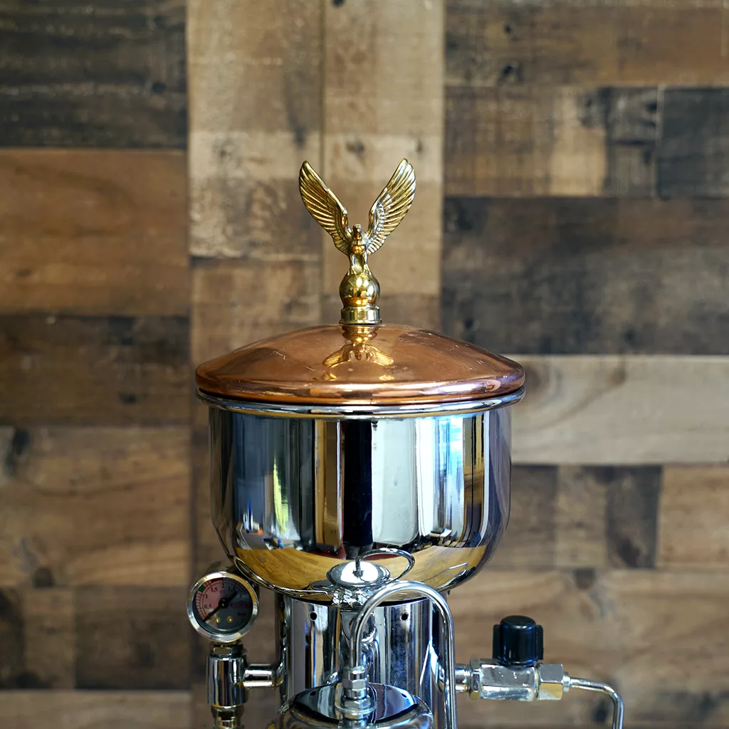 The SIDEWALK SALE - Elektra SXC Microcasa Semiautomatica Chrome Espresso Machine USED (E211) features a shiny chrome body and a copper lid with a golden eagle figurine, displayed against a wooden panel wall. © 2025 1st-line.com