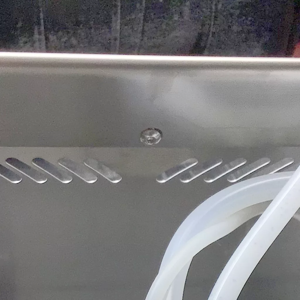 A close-up of a Cuadra metallic surface with diagonal slits, a small screw, and translucent flexible tubing coiled at the bottom right corner, hinting at accessing the inside. The background appears slightly blurry and dark. © 2025 1st-line.com