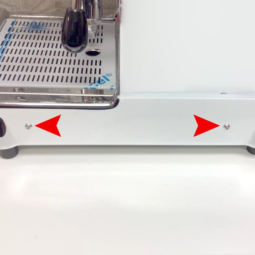 A close-up of the bottom front panel of a coffee machine inside Cuadra, with two red arrows pointing to screws on each side. © 2025 1st-line.com