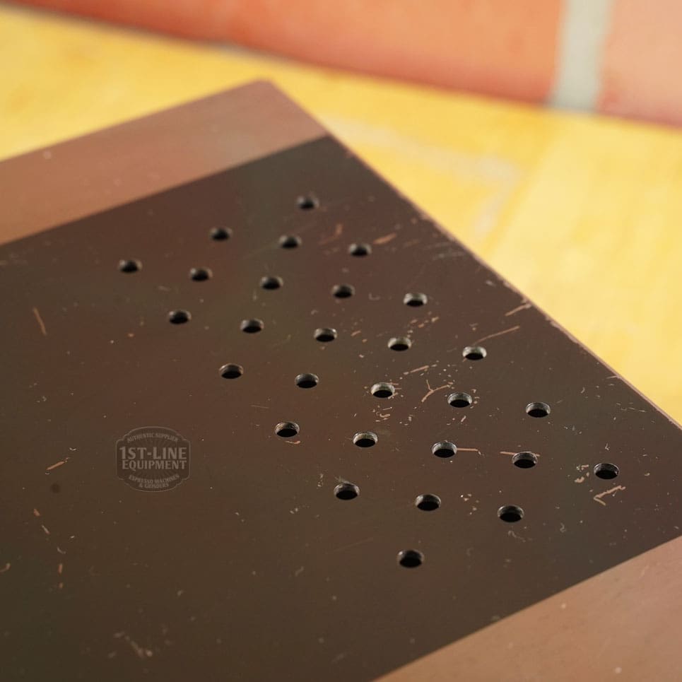 A close-up of the SIDEWALK SALE - Lelit PLA360M Knock Box Drawer (E208) shows its brown metal surface with rows of small round holes, light scratches, and a slightly reflective finish against a blurred wood and brick background. © 2025 1st-line.com