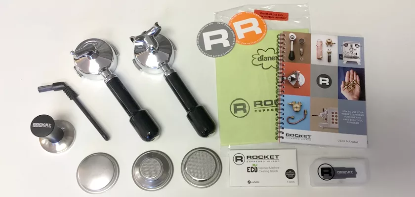 A Rocket Mozzafiato Cronometro Type V HX Espresso Machine accessory kit—portafilters, metal baskets, tamper, brush, manuals, stickers, and warranty card—is neatly arranged on a white surface. © 2025 1st-line.com