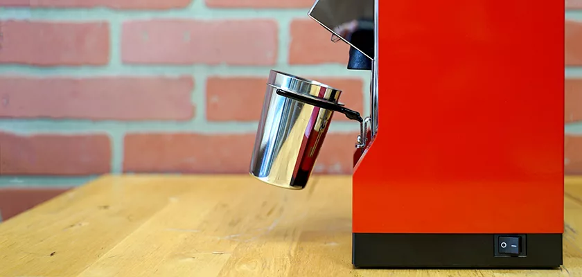 A shiny metal cup attached to a red espresso machine sits on a wooden table with a brick wall in the background. © 2025 1st-line.com