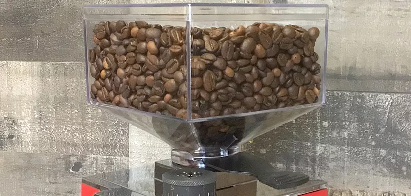 A transparent hopper of dark roasted coffee beans sits atop the Eureka Mignon Libra Espresso Coffee Grinder - Weight-Based Grinding, set against a textured gray wall. © 2025 1st-line.com