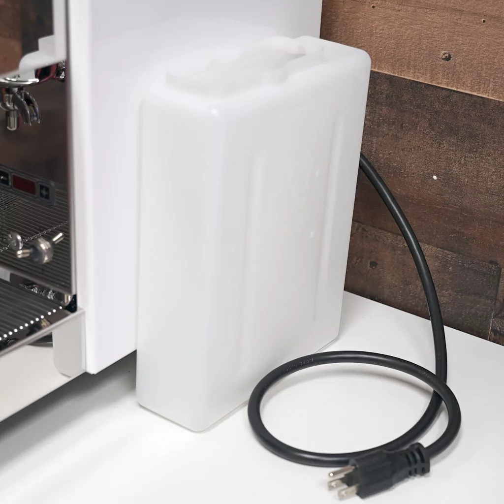 A white plastic water reservoir attached to the back of a Bezzera Crema coffee machine sits on a white surface, with a black power cord coiled nearby. A brown wood panel is in the background. © 2025 1st-line.com