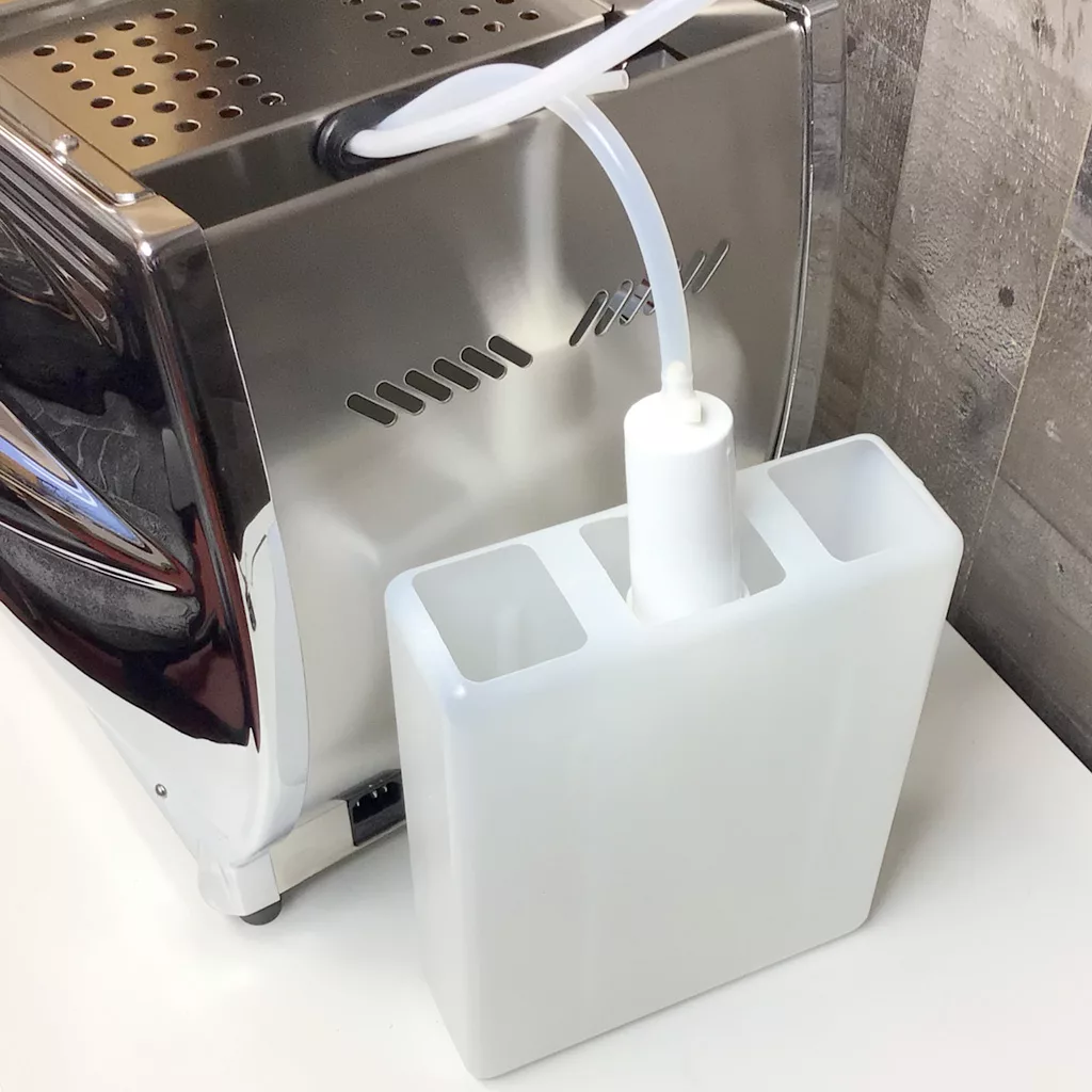 A white plastic water tank with attached tubing is connected to the back of a stainless steel Cuadra espresso machine, sitting on a white countertop near a gray wall, with easy access for accessing the inside. © 2025 1st-line.com