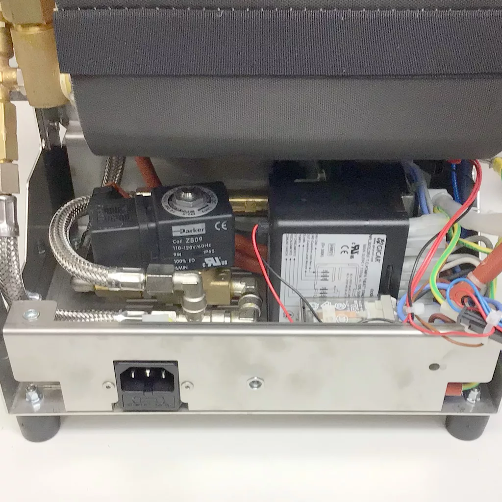 Accessing the inside of the Cuadra, the image reveals internal components such as electrical wiring, switches, a solenoid valve, metal pipes, and a visible power input socket on a metallic base. © 2025 1st-line.com