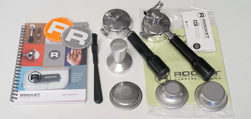 Accessories for the Rocket Appartamento TCA HX Espresso Machine—including user manual, metal tamper, cleaning brush, two portafilters, filter baskets, Rocket stickers, cloth, and ECO info card—arranged on a white surface. © 2025 1st-line.com
