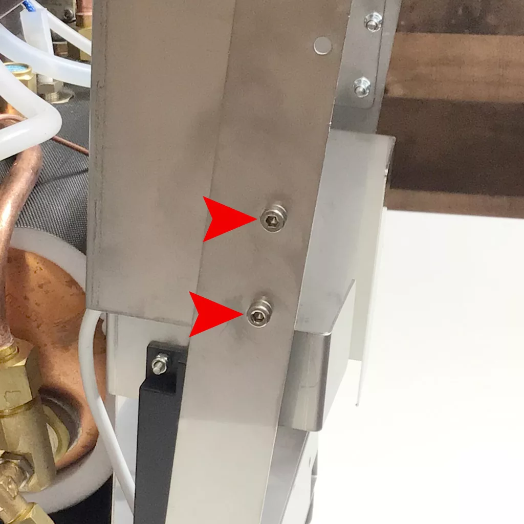 Close-up of a Cuadra metal panel with two red arrows pointing at screws, indicating points for accessing the inside. Copper and plastic tubing, along with other mechanical components, are partially visible in the background. © 2025 1st-line.com