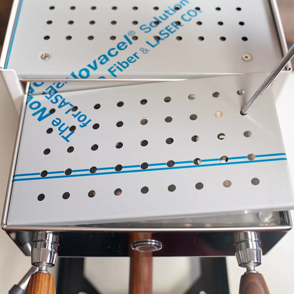 Close-up of a metallic, perforated tray with blue text and lines—possibly part of DE PM medical or laboratory equipment—with wooden-handled tools visible at the bottom, offering a glimpse of inside access. © 2025 1st-line.com