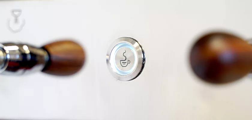 Close-up of a round button with a coffee cup icon, bordered by two espresso machine handles with wooden grips on either side, against a white background. © 2025 1st-line.com