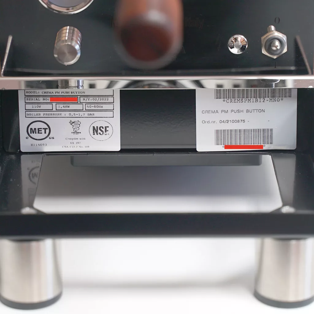 Close-up of Bezzera Crema PM coffee machine identification labels, including model, voltage, and certification details attached below the control panel, with a reflection visible on the metal drip tray underneath. © 2025 1st-line.com