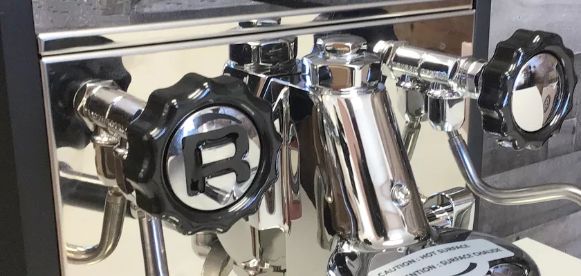 Close-up of shiny chrome Rocket Mozzafiato Cronometro Type V HX Espresso Machine parts, featuring black handles with a distinctive R logo and various knobs and levers reflecting the environment. © 2025 1st-line.com