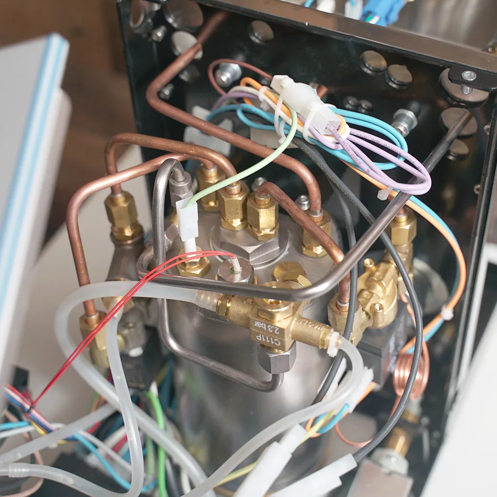 Close-up of the internal components of a Bezzera Crema PM espresso machine, showing metal pipes, brass fittings, colorful electrical wires, and tubes connected to a cylindrical metal boiler. © 2025 1st-line.com