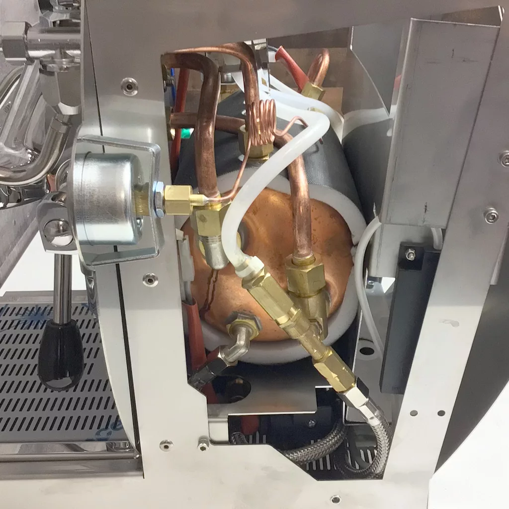 Close-up of the internal components of a Cuadra espresso machine, showing copper pipes, a boiler, brass fittings, and various connecting tubes and wires inside a metallic housing after accessing the inside. © 2025 1st-line.com