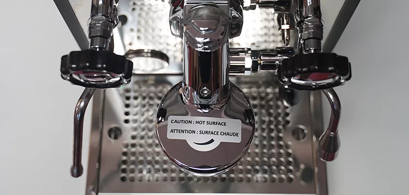 Close-up of the Rocket Mozzafiato Cronometro Type V HX Espresso Machine group head with a Caution: Hot Surface / Attention : Surface Chaude warning label on the front and a metal drip tray beneath. © 2025 1st-line.com