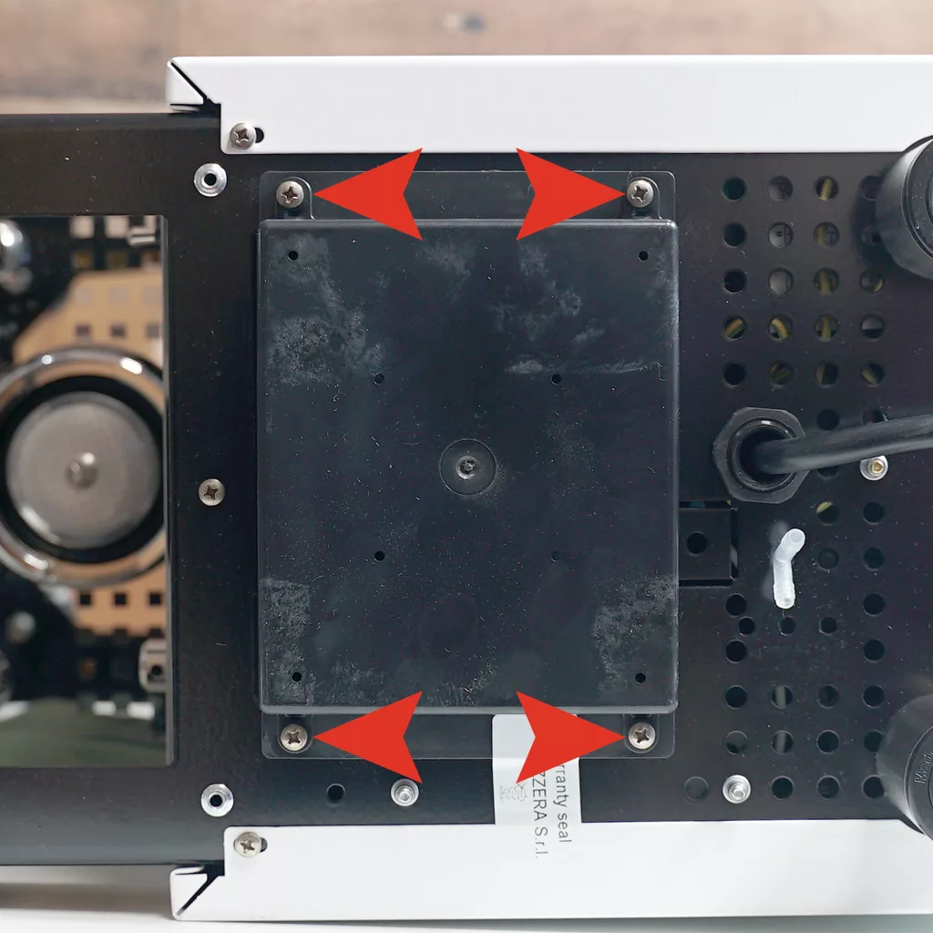 Close-up of the underside of a Bezzera Crema DE with four red arrows pointing at screws on the corners of a black rectangular panel, indicating where the panel is secured to the device. © 2025 1st-line.com