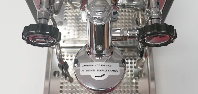 Close-up top view of a shiny Rocket Appartamento TCA HX Espresso Machine featuring metal components, black knobs, a perforated drip tray, and a CAUTION: HOT SURFACE sticker in both English and French. © 2025 1st-line.com