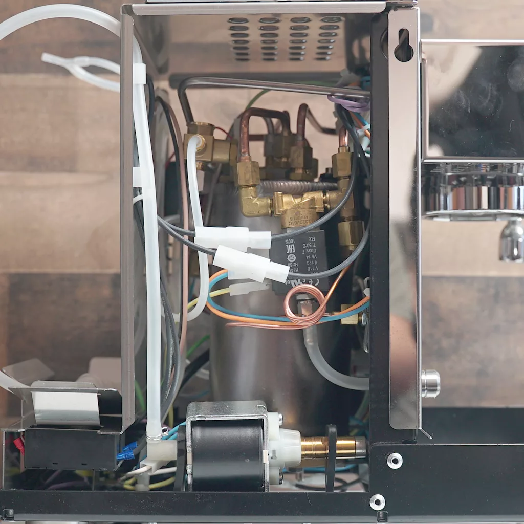 Close-up view of the internal components of a Bezzera Crema espresso machine, showing various wires, metal pipes, and a pump inside a partially open stainless steel casing. © 2025 1st-line.com