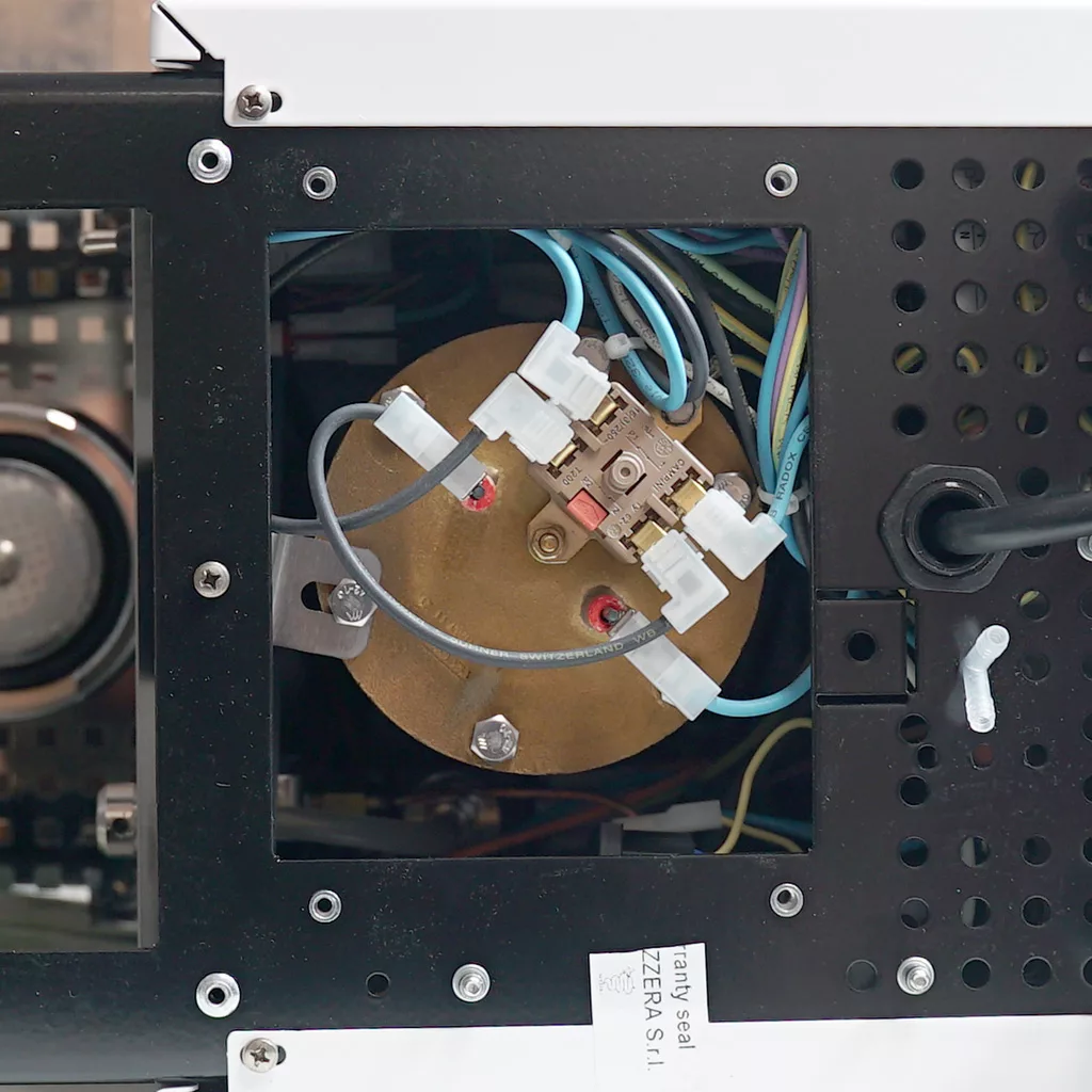 Close-up view of the internal wiring and components of a Bezzera Crema espresso machine, showing inside access to a round brass part with connected wires, plastic connectors, and a black perforated metal casing. © 2025 1st-line.com