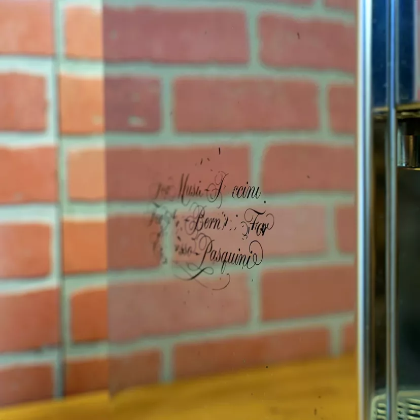 A close-up of a mirrored surface reflecting handwritten cursive text, with a red brick wall and a (SOLD) SIDEWALK SALE - Pasquini Livia G4 PID Electronic Espresso Machine (USED) in the background, adding vintage flair to the modern scene. © 2025 1st-line.com