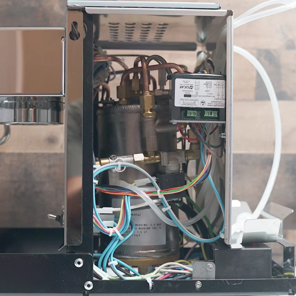 Side view of an open Bezzera Crema espresso machine showing internal components, including wires, a metal boiler, copper pipes, a relay module, and connectors; background features a wooden surface. © 2025 1st-line.com