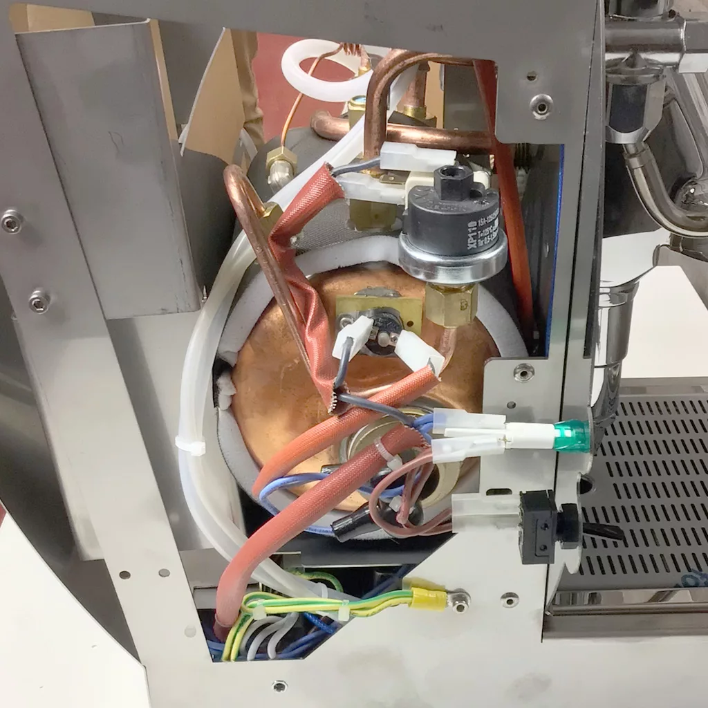 The image shows the internal components of a Cuadra espresso machine, including copper tubes, wires, a pressure valve, and a large cylindrical boiler, all visible when accessing the inside through its metallic frame. © 2025 1st-line.com