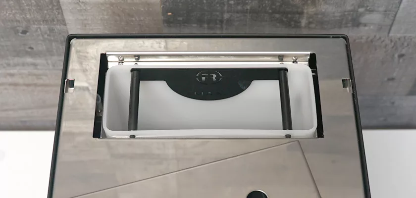 Top-down view of a rectangular opening with a flap on the Rocket Appartamento TCA HX Espresso Machine, positioned against a wood-patterned background. © 2025 1st-line.com