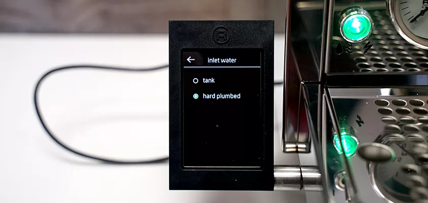 A close-up of a coffee machine display screen showing water inlet options: “tank” and “hard plumbed.” The machine has metal parts and green indicator lights. A power cord is visible in the background. © 2025 1st-line.com