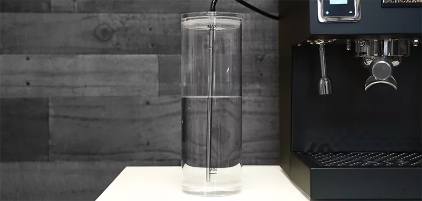 A transparent water reservoir sits next to the Bellezza Bellona Dual Boiler Espresso Machine on a white countertop, with a grey wood-patterned wall in the background. © 2025 1st-line.com