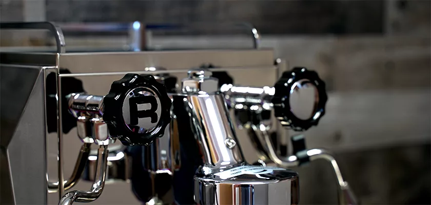 Close-up of a shiny Rocket Giotto Cronometro Type V HX Espresso Machine with stainless steel finish and black knobs, including one marked R, set against a blurred wooden background. © 2025 1st-line.com