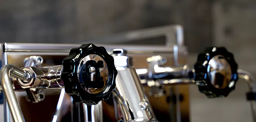 Close-up of two black, ridged knobs on a shiny, metallic espresso machine with blurred, neutral-colored background. © 2025 1st-line.com