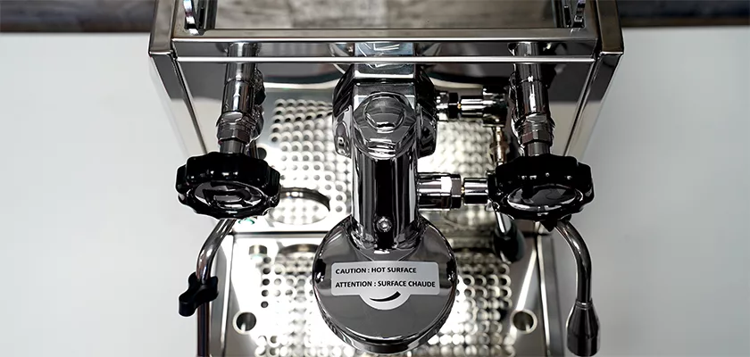 Top view of the Rocket Giotto Cronometro Type V HX Espresso Machine, featuring two black knobs, a central portafilter holder, a frothing wand, and a “CAUTION: HOT SURFACE” warning label in English and French. © 2025 1st-line.com