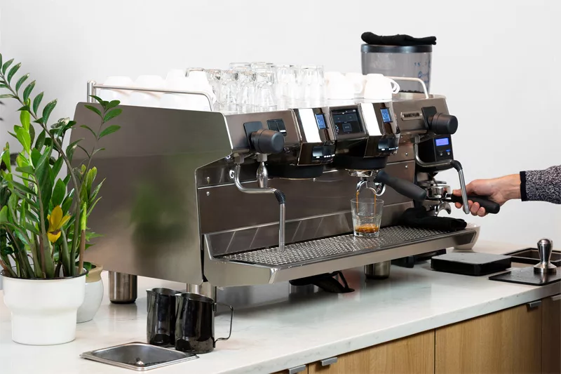 A person uses the Rancilio Invicta 2 Group Commercial Espresso Machine on a countertop, with glass cups placed on top, a potted plant nearby, and a small glass collecting espresso under the portafilter. © 2025 1st-line.com